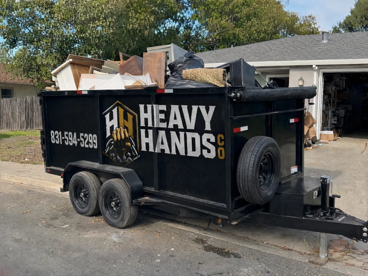 Heavy Hands Co. dump trailer loaded with cabinets, lumber and bagged debris outside an open garage during a Salinas cleanout